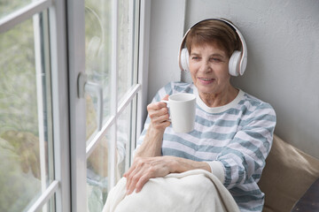  Audio healing. concept without stress. senior woman in headphones listens to music, drinks tea at home 