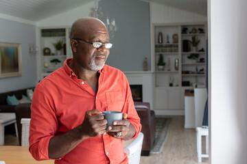 Thoughtful african american senior man having coffee while standing in living room at home