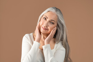 Lovely senior woman with beautiful long grey hair touching her face over brown studio background