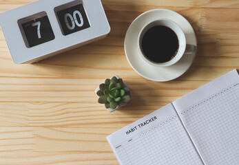 flat lay of habit tracker book, a cup of black coffee, flip clock 7 am and succulent plant pot on wooden table background with copy space.