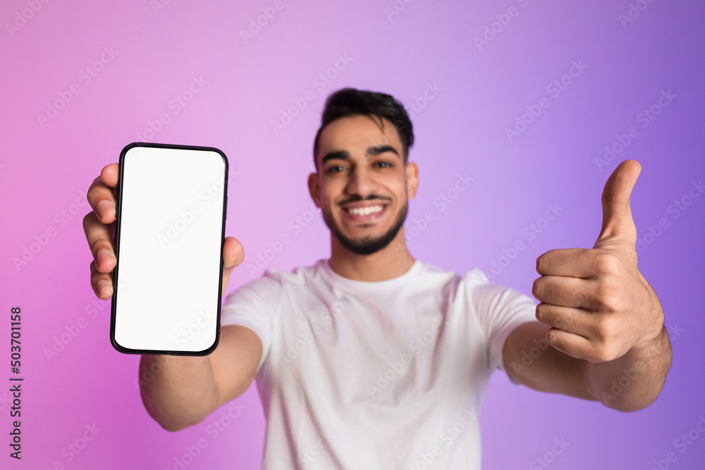 Poster happy yougn arab guy holding cellphone with empty white screen and showing thumb up gesture in neon 