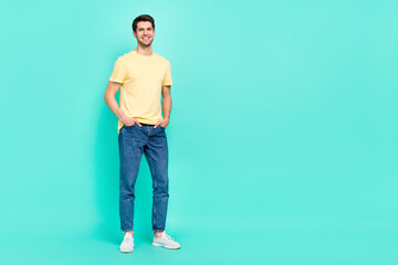 Fototapeta na wymiar Full size photo of cute brunet young guy near empty space wear t-shirt jeans shoes isolated on turquoise color background