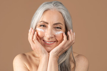 Mature beauty. Charming senior woman posing with face cream under her eyes, smiling at camera over...