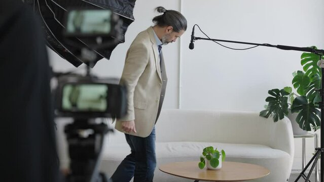 Male Business Man Walks Into Video Production Set and Sits Down Ready to be Interviewed. Behind the Scenes Camera, Lighting and Audio Equipment Can Be Seen. Slow Motion Scene of On Camera Talent