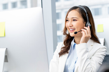 Cheerful Asian woman worker wearing headset with microphone talking to client while working in...