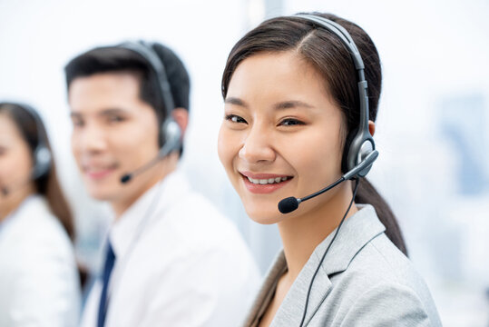 Optimistic smiling Asian female in headset with microphone looking at camera while working in call center with coworkers