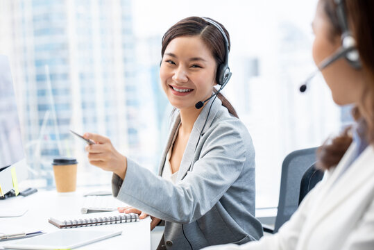 Positive Asian female adviser wearing headset with microphone looking at colleague while sitting at table in modern office
