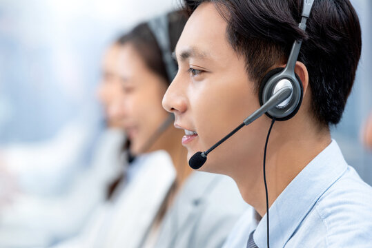 Side view of Asian man consultant in headset with microphone working in modern call center with colleagues on blurred background