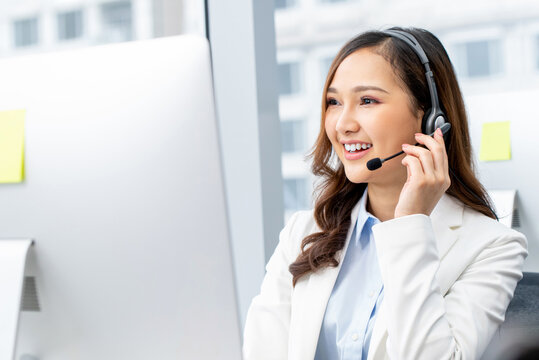 Cheerful Asian woman worker wearing headset with microphone talking to client while working in mordern call center office