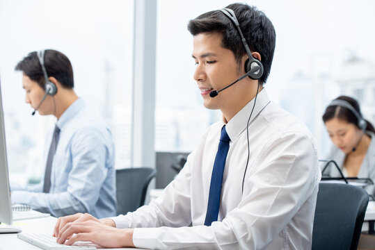 Side view of focused Asian male worker in headset typing on computer while working in call center with coworkers