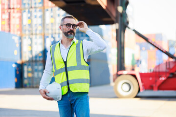 Handsome caucasian male dock foreman worker in white hard hat helmet and high-visibility vest working in Shipping Cargo Container Terminal Depot