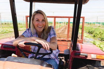 Portrait of happy caucasian mid adult female farmer with blond hair sitting in tractor at greenhouse