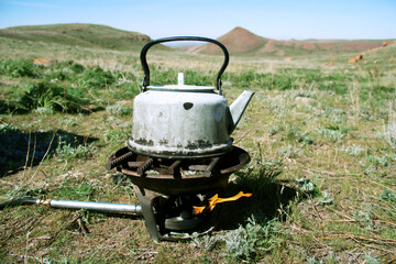 An old smoky white camping kettle on a gas burner in the mountains near the Malaysary gorge, in the distance a green plateau and hills against a clear sky, summer, sunny