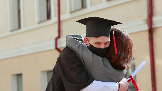 Mother in face mask hugs her son student in graduation gown and a square cap