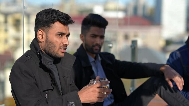 Side View Relaxed Young Men Sitting In Sunbeam On Rooftop As Unrecognizable Waitress Bringing Alcohol Glasses. Carefree Middle Eastern Friends Partying Outdoors With Whiskey On Sunny Day