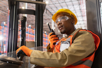 Low angle view of african american young male worker sitting in forklift talking over walkie-talkie