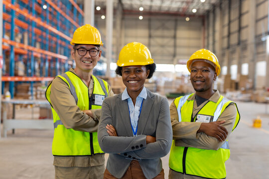 Smiling multiracial female and male coworkers wearing hardhats with arms crossed in warehouse