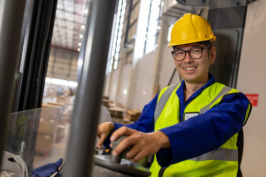Portrait of smiling asian mature male worker wearing reflective vest and hardhat driving forklift