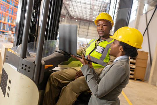 African american young female foreman showing laptop to young male coworker sitting in forklift