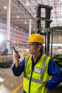 Asian mature male warehouse worker wearing reflective vest and hardhat talking over walkie-talkie