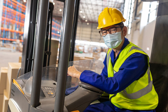 Portrait of asian mature male worker wearing mask, reflective vest and helmet while driving forklift