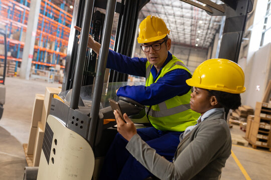 African american young woman discussing over digital pc with asian male coworker sitting in forklift