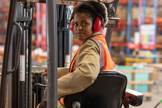 African american young female worker wearing ear protectors looking away while sitting in forklift
