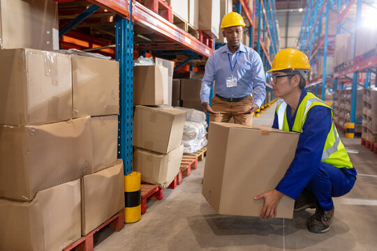 African american young foreman guiding asian mature male worker while lifting cardboard box by shelf