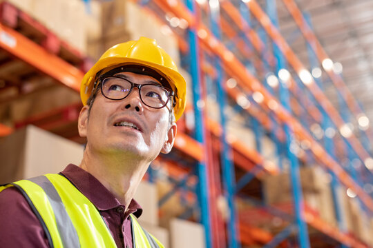 Low angle view of asian mature male warehouse worker wearing hardhat and eyeglasses looking away