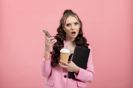 Portrait of shocked, puzzled girl in jacket reading post on social networking site using phone, chatting, holding laptop and coffee mug looking puzzled. Studio shot isolated on pink background.