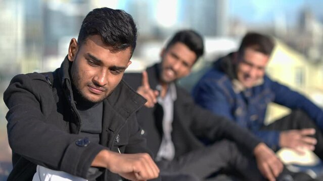 Young sad Middle Eastern man looking at camera as blurred friends laughing pointing mocking at background. Portrait of bullied handsome guy posing on sunny day outdoors on rooftop. Lifestyle