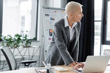 stylish economist standing at workplace, using laptop and looking away.