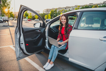 Portrait of attractive cheerful skilled wavy-haired girl sitting in auto writing email service order on parking outdoors