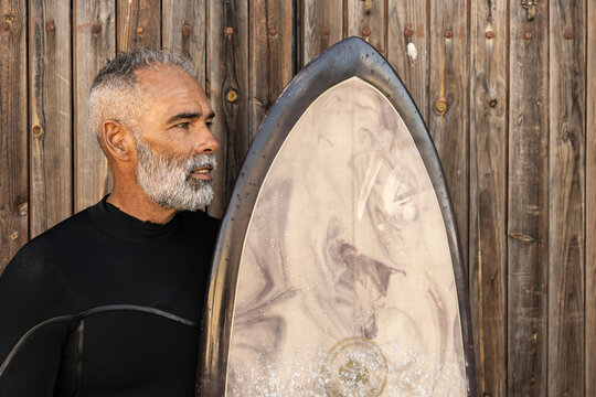 A Middle-aged African Man In A Surfing Costume Holding His Surfing Board And Looking Aside