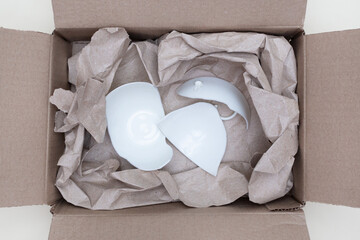 Broken white cup in a box with packing paper for shipping. Flat lay. Top view. Concept of careless...