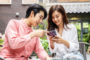 Happy Young asian woman sitting with boyfriend at cafe table, discussing over on smartphone, Young couple looking at mobile phone shopping online and share information together in coffee shop