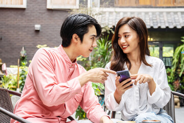 Happy Young asian woman sitting with boyfriend at cafe table, discussing over on smartphone, Young couple looking at mobile phone shopping online and share information together in coffee shop