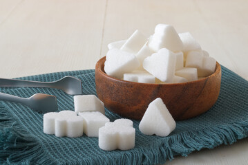 Bridge-shaped sugar cubes in wooden bowl on a turquoise napkin with silver sugar tongs. Front closeup view, white wooden table, no people.