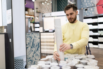 Obraz na płótnie Canvas a man in a store of decorative building finishing materials chooses paper wallpaper for renovation in an apartment