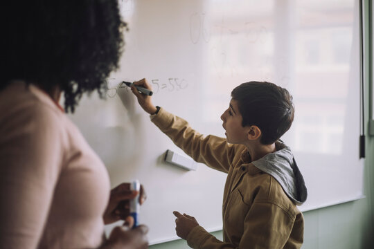 Side view of boy solving maths sum on white board with teacher in classroom