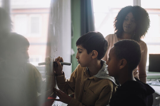 Boys doing mathematics on white board while teacher examining in classroom