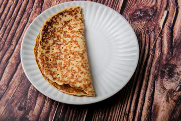 Plain sweet pancake folded in half on a small white plate against a wooden background.