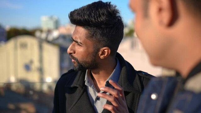 Confident young man looking away listening and talking to blurred friend standing at front. Portrait of Middle Eastern guy chatting with mates on rooftop in urban city on sunny day