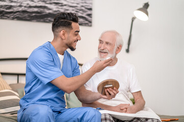 Cheerful caregiver preparing an aged male person for a shave
