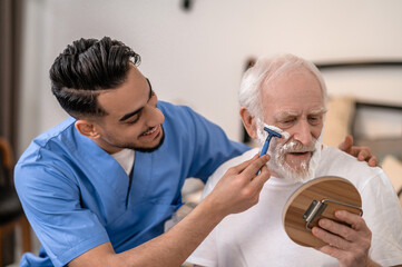 Joyous young man shaving a serious old gentleman