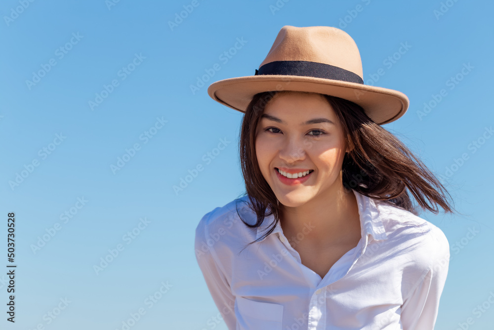 Wall mural Portrait happy attractive young Asian woman wear hat looking at camera in the bright sunny day summer blue sky. Millennial lady with smiley face on her vacation holiday trip in tropical location