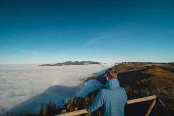 Fototapeta na wymiar tourist watches the highest rocky mountains of the island of Madeira emerge from the clouds into the sunset from the viewpoint of Pico Ruivo do Paul. Beauty of the Portuguese island. Above the clouds
