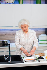 An old well-groomed blonde woman cooks a trout steak in the kitchen