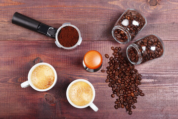 Two cups of coffee, holder with ground coffee, tamper and cans of coffee beans on wooden table. View from above.