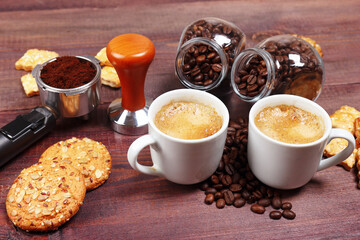 Two cups of espresso, crackers, holder with ground coffee, tamper and coffee beans on wooden table.